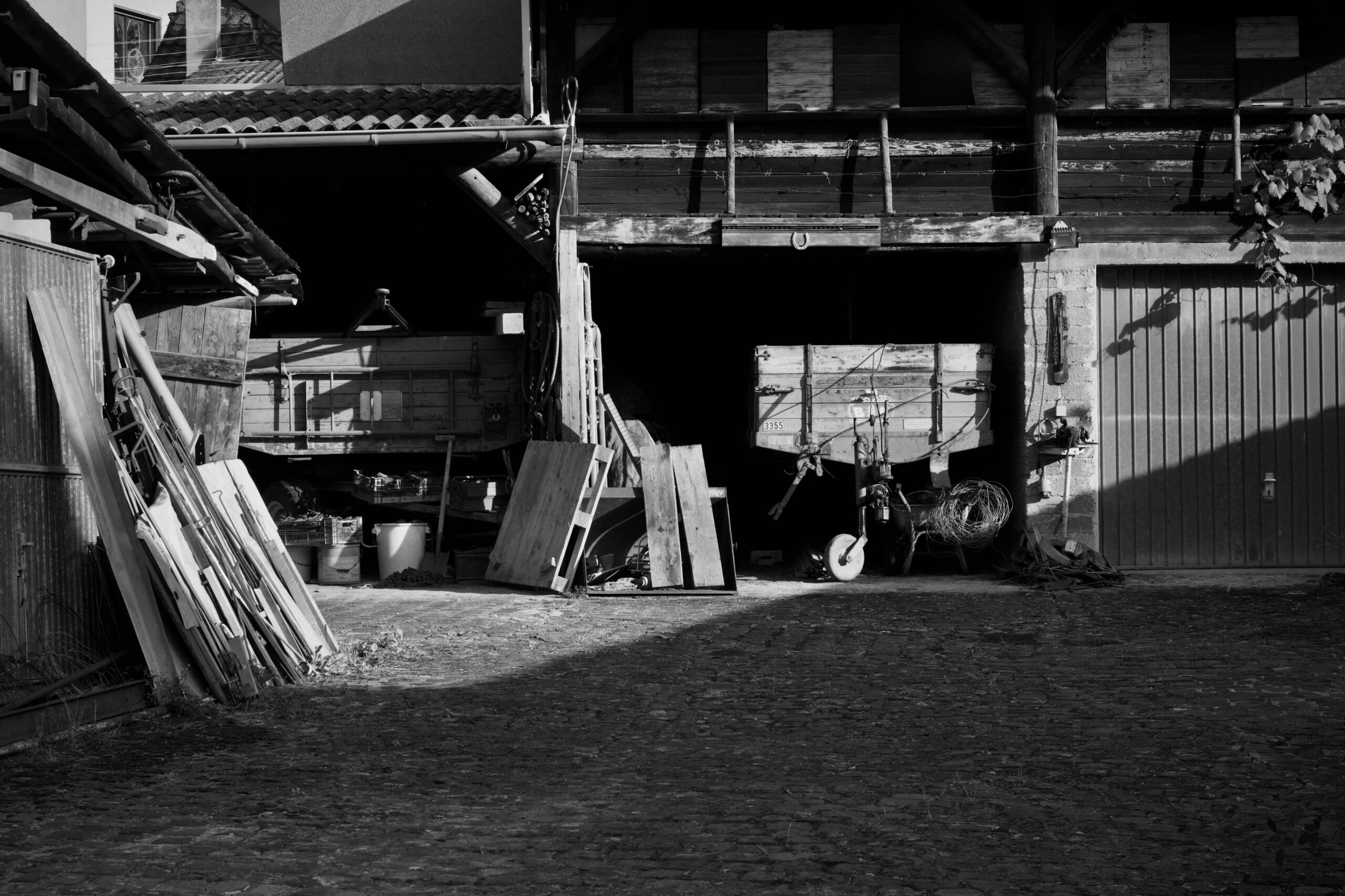 Ein Schwarzweißfoto, das einen rustikalen Scheunenhof mit alten Holzkonstruktionen und Schatten zeigt.