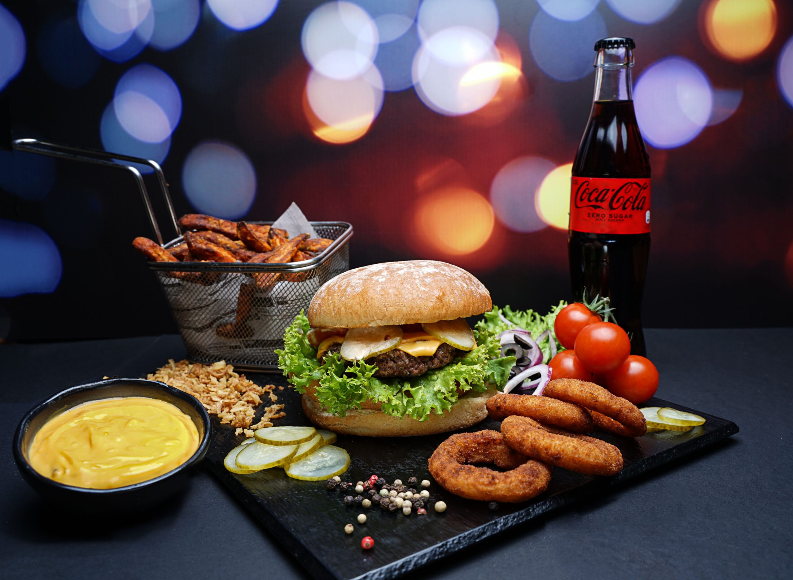 Ein Gourmet-Burger mit Beilagen, Soße und Cola vor einem lebendigen Bokeh-Hintergrund.